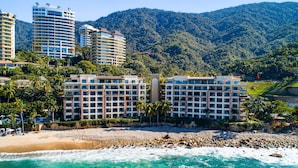 Un hotel frente a la playa con varios pisos y montañas al fondo.