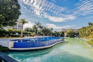 A swimming pool with a tiled edge, surrounded by greenery and palm trees, with buildings in the background. - Golf Course-adjacent Getaway With Infinity Pool, Private Plunge Pool & Balcony (Nuevo Nayarit)
