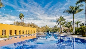Ein Pool mit klarem, blauem Wasser, umgeben von Palmen und einem gelben Gebäude mit Bogenfenstern.