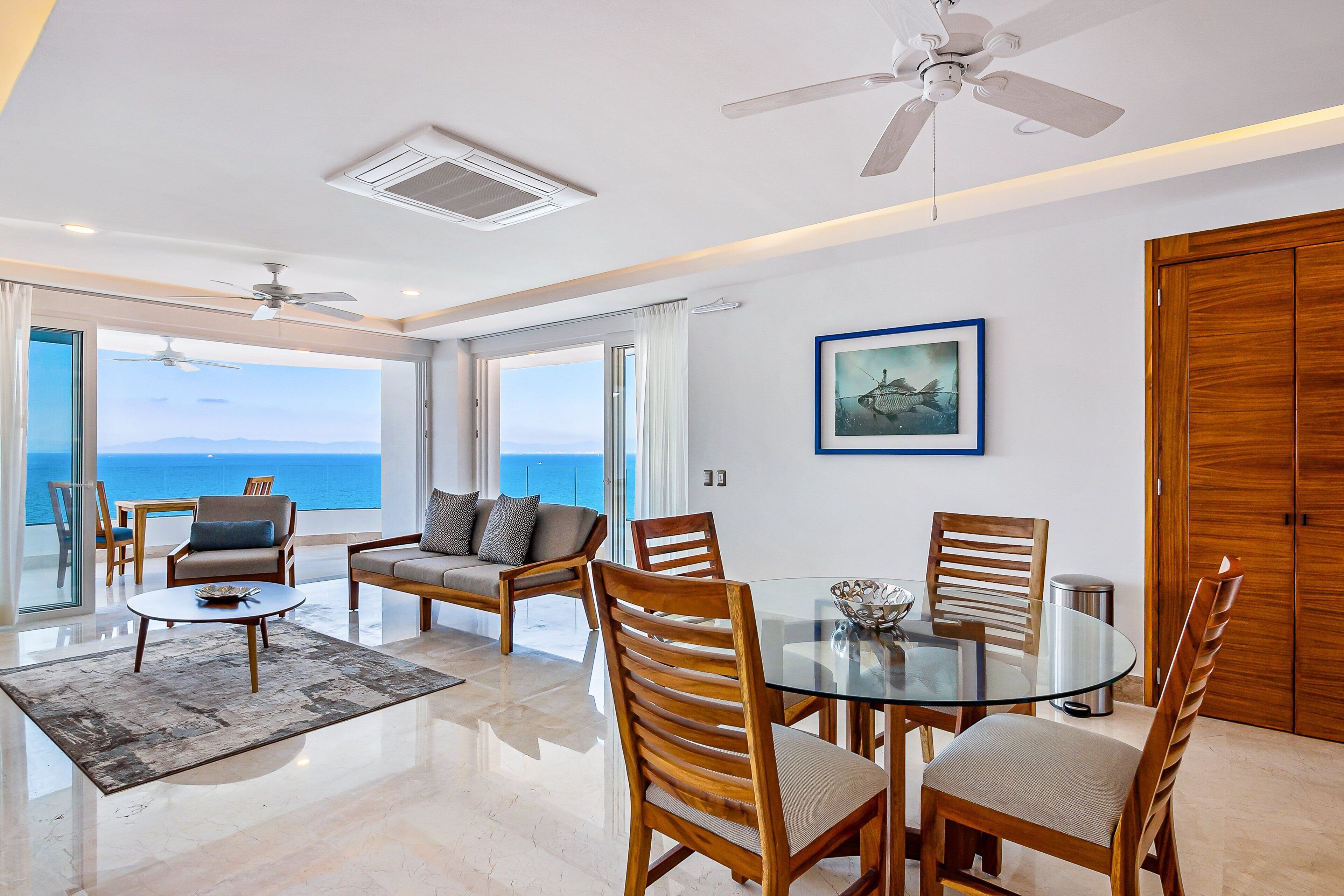 A spacious living area with a glass table, wooden chairs, a sofa, and a ceiling fan.