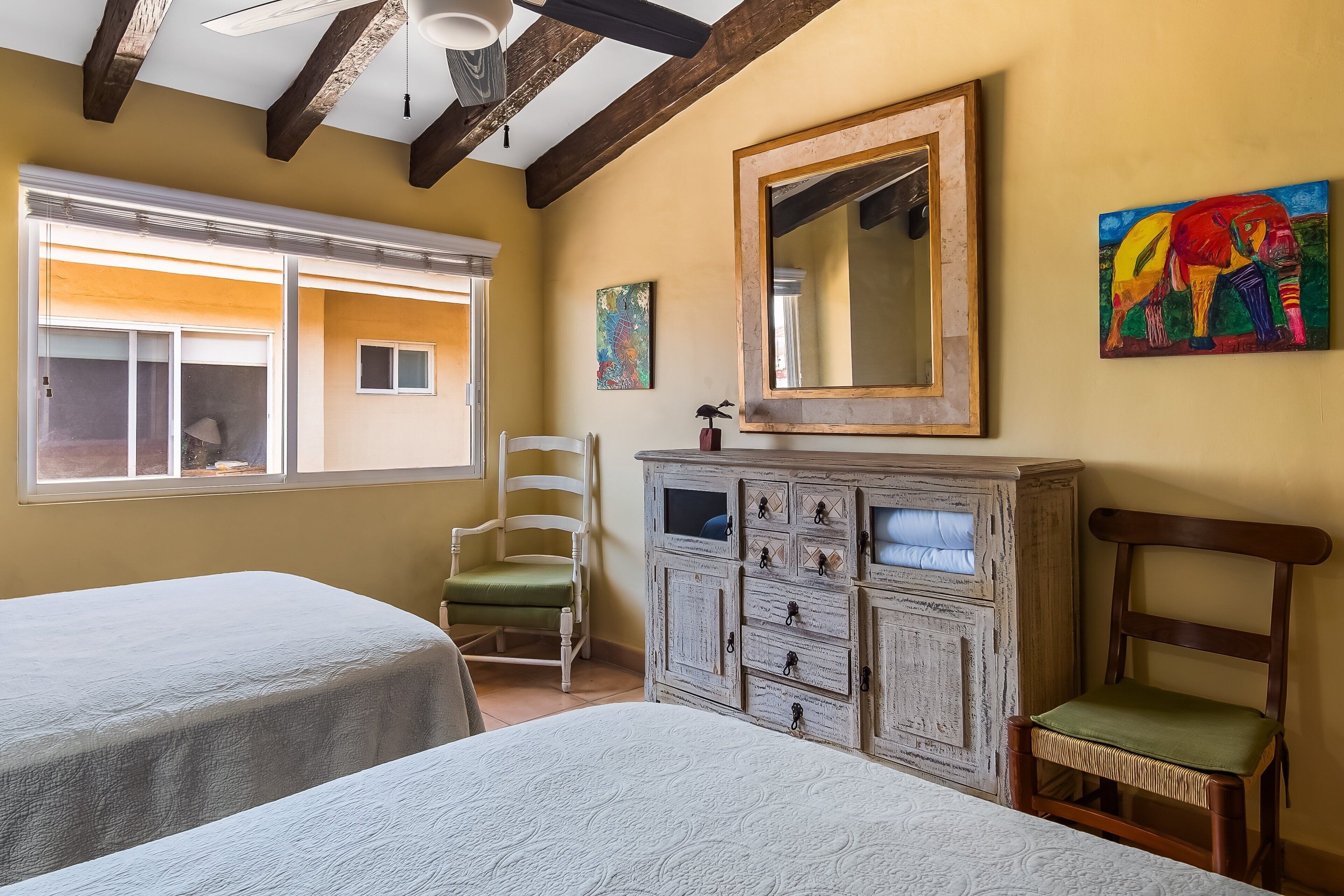 A room with two beds, a wooden cabinet, a chair, a window, and a painting.