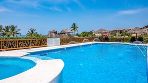 A swimming pool with a curved edge, palm trees, and thatched-roof structures. - Oceanfront Condo With Pool, Balcony, Amazing Views, Washer/dryer, & Partial AC (Punta de Mita)