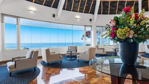 A spacious lobby with a large glass window offering a view of the ocean, a seating area with chairs, a round table, and a large vase of flowers. - Upscale Coastal Suite With Private Terrace, Ocean Views, Pool & hot tub (Puerto Vallarta)