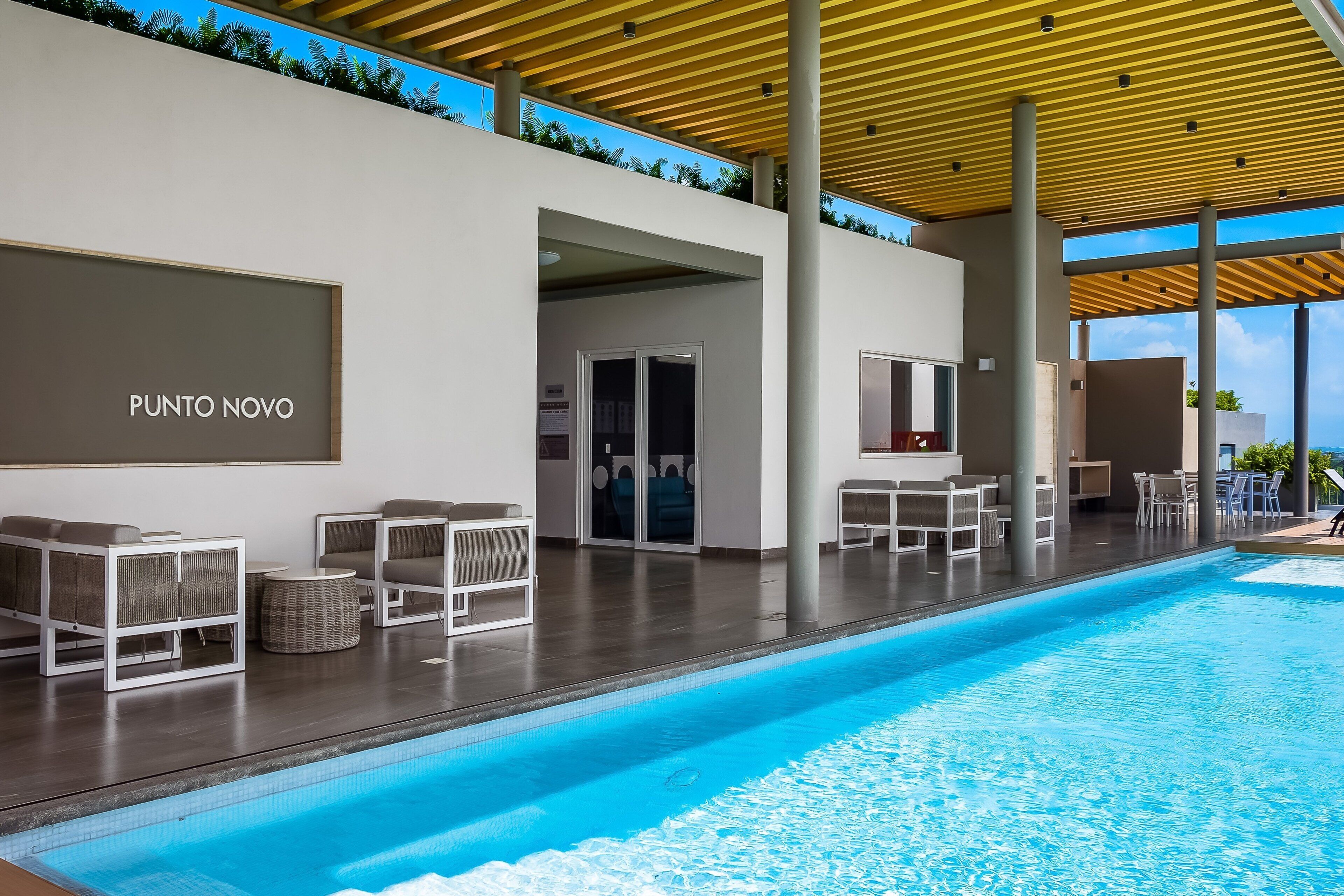 Un área de piscina moderna con sillones de descanso, una pérgola de madera y un letrero que dice 'PUNTO NOVO'.