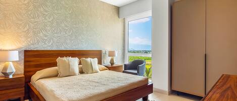A bedroom with a large bed, nightstands, a chair, a wardrobe, and a view of greenery.