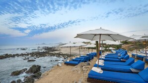 Een terras aan het strand met blauwe ligstoelen en witte parasols, met uitzicht op de oceaan en de rotsachtige kustlijn.