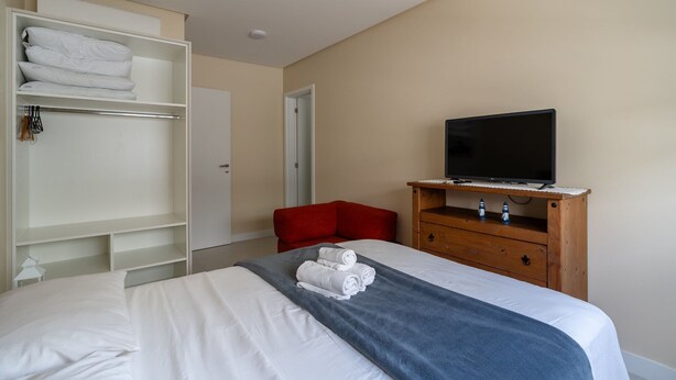 Quarto de hotel com uma cama, televisão sobre um suporte de madeira, uma cadeira vermelha e um guarda-roupa com toalhas dobradas.
