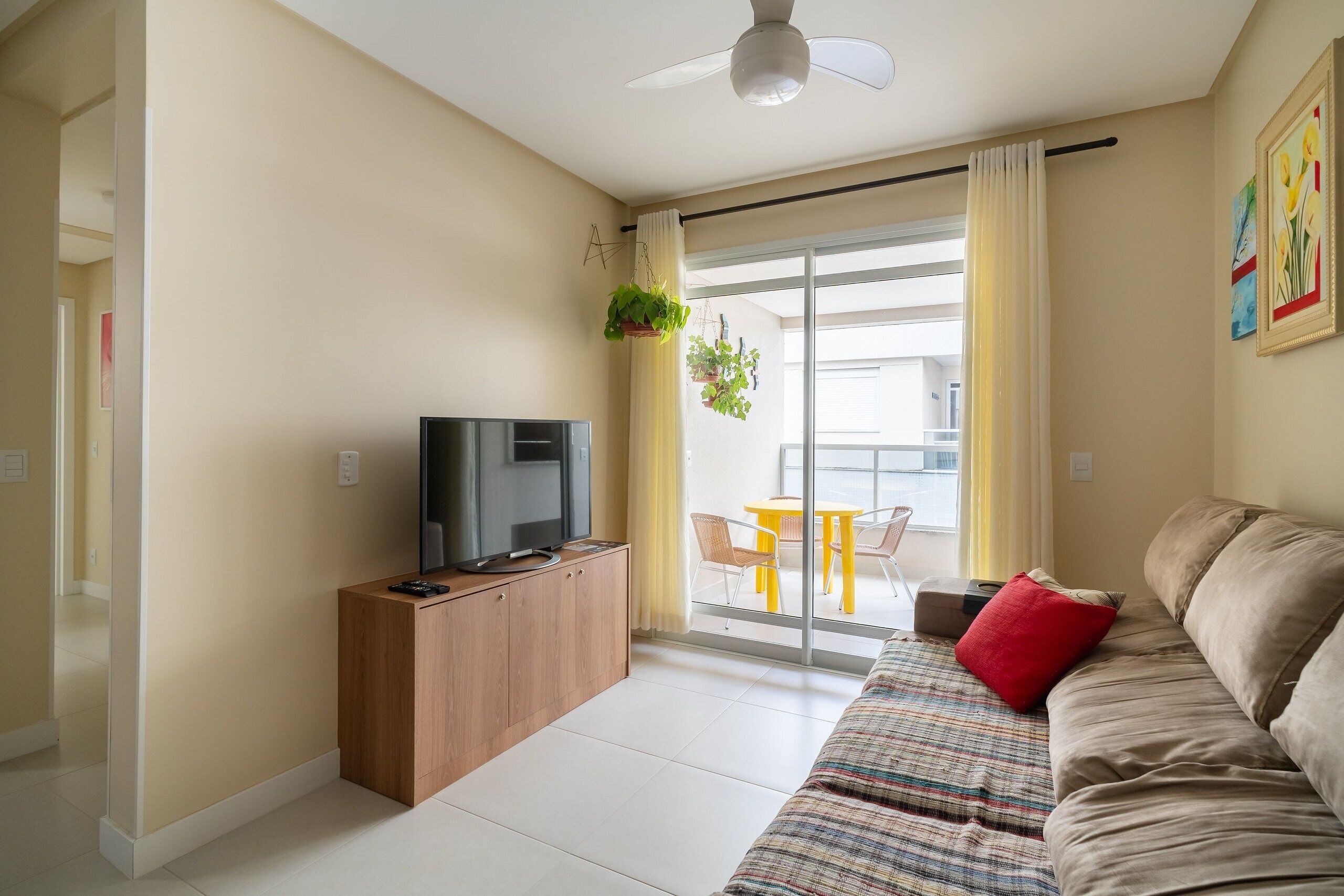 A living room with a sofa, a television, a wooden cabinet, a balcony with yellow chairs, and a sliding glass door.