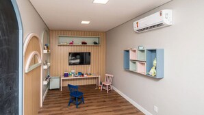 Quarto com piso de madeira, uma escrivaninha, cadeiras, prateleiras com brinquedos e um aparelho de ar condicionado.