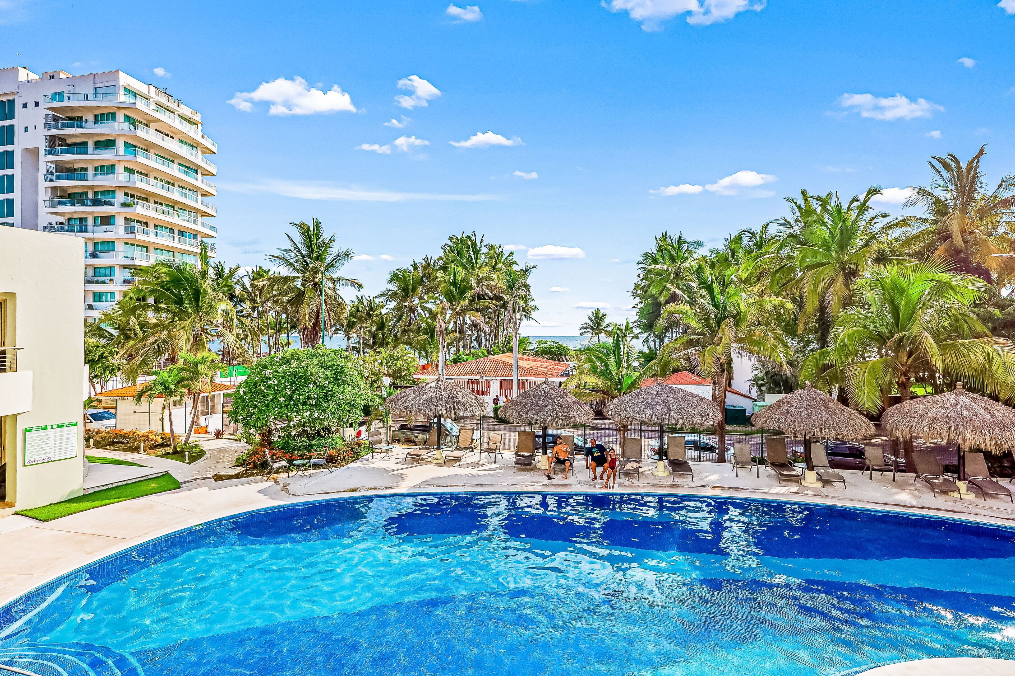 Piscina com espreguiçadeiras e palmeiras, um edifício de vários andares e um céu azul límpido.
