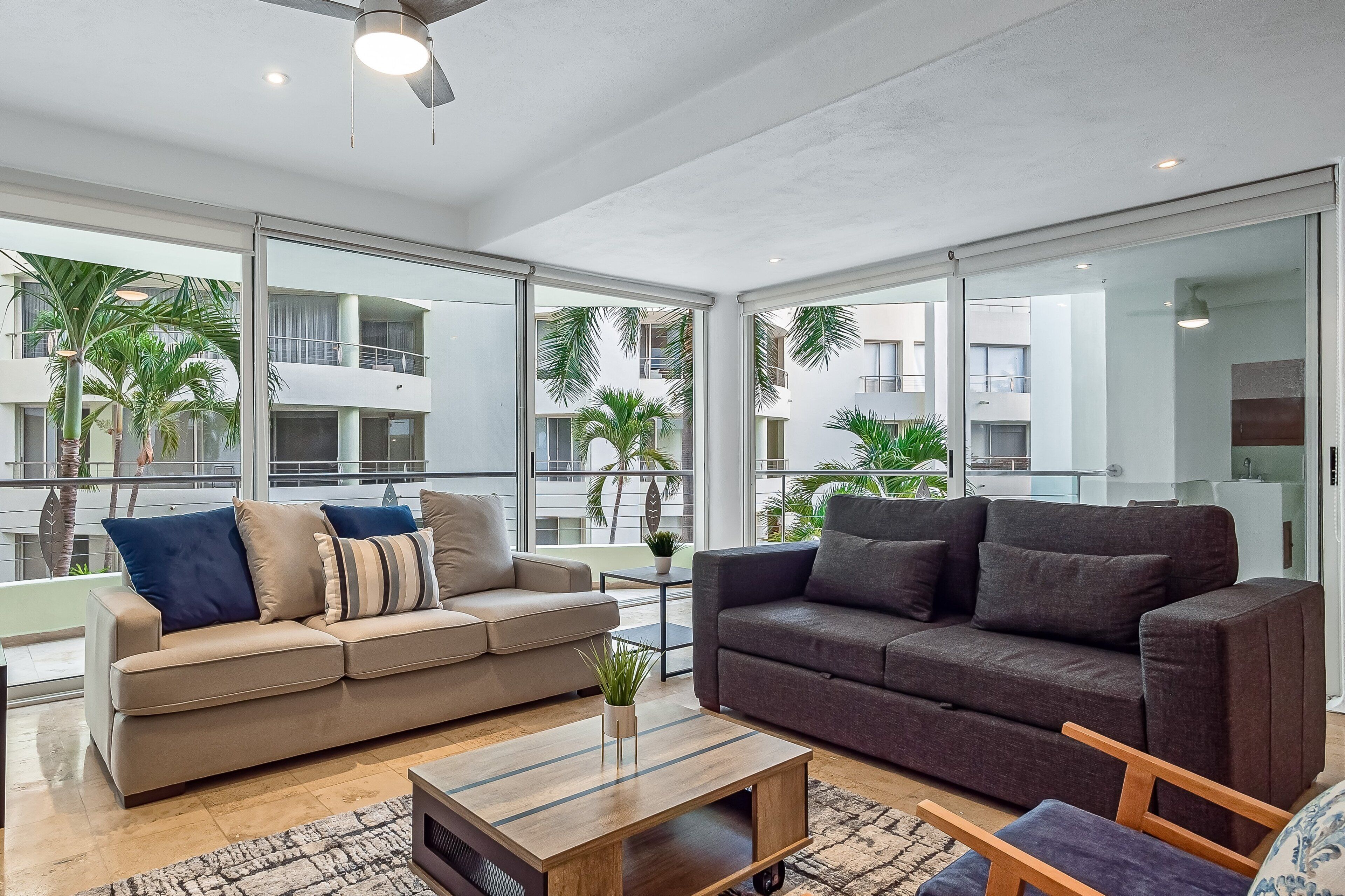 A modern living room with two sofas, a coffee table, and a view of a residential area with balconies and palm trees.