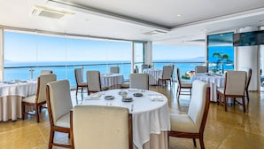 A dining area with round tables, chairs, and a large window overlooking the sea. - Stylish Hillside Suite W/rooftop Pool, gym Access, AC & Family-friendly Layout (Puerto Vallarta)