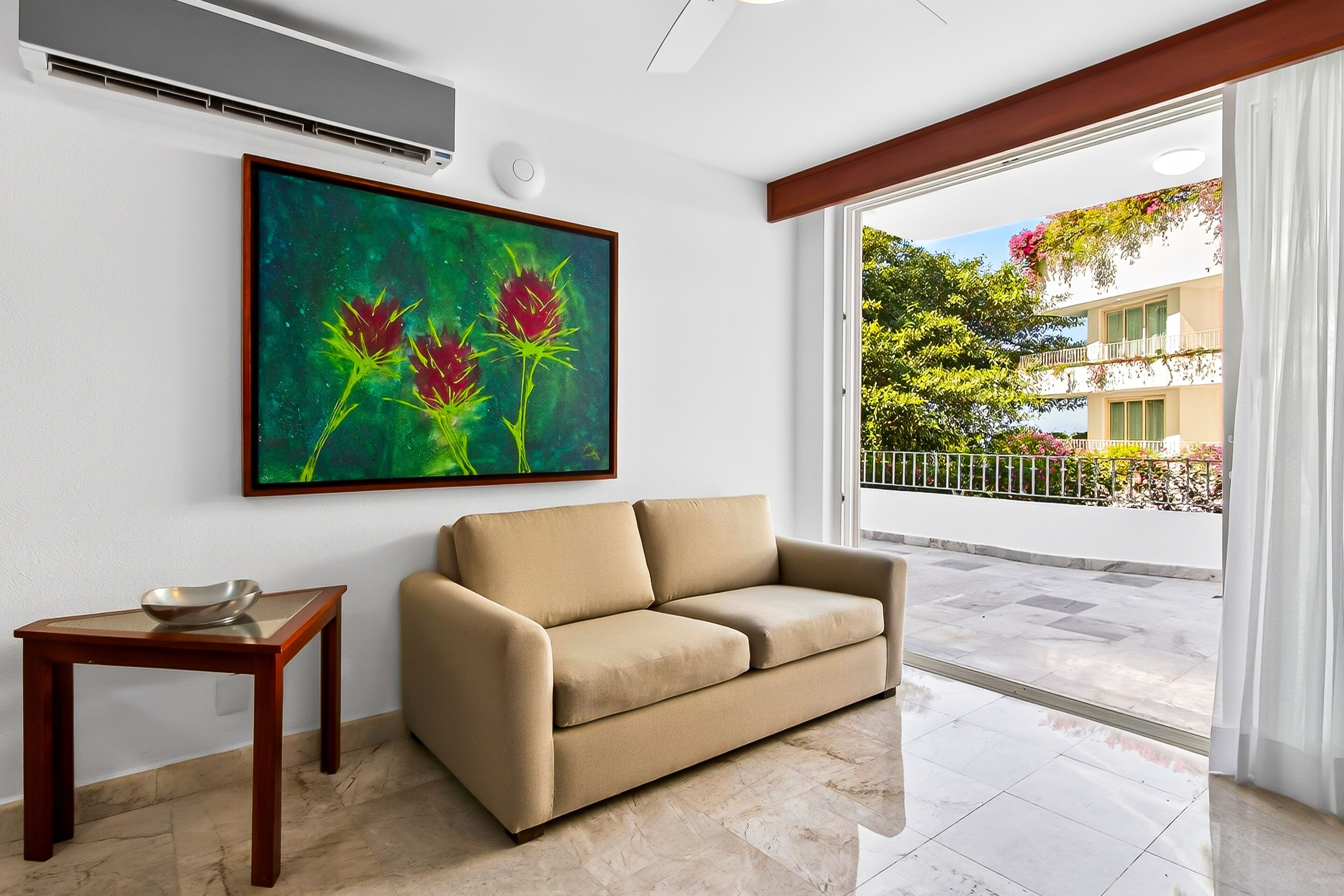 Un salón con un sofá color beige, una pequeña mesa de madera con un tazón, un cuadro abstracto grande y vistas a un balcón con plantas y un edificio.