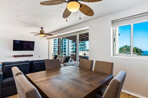 Een moderne woonkamer met een houten eettafel, comfortabele zithoek, een plafondventilator, een flatscreen televisie en grote ramen met uitzicht op zee en gebouwen.