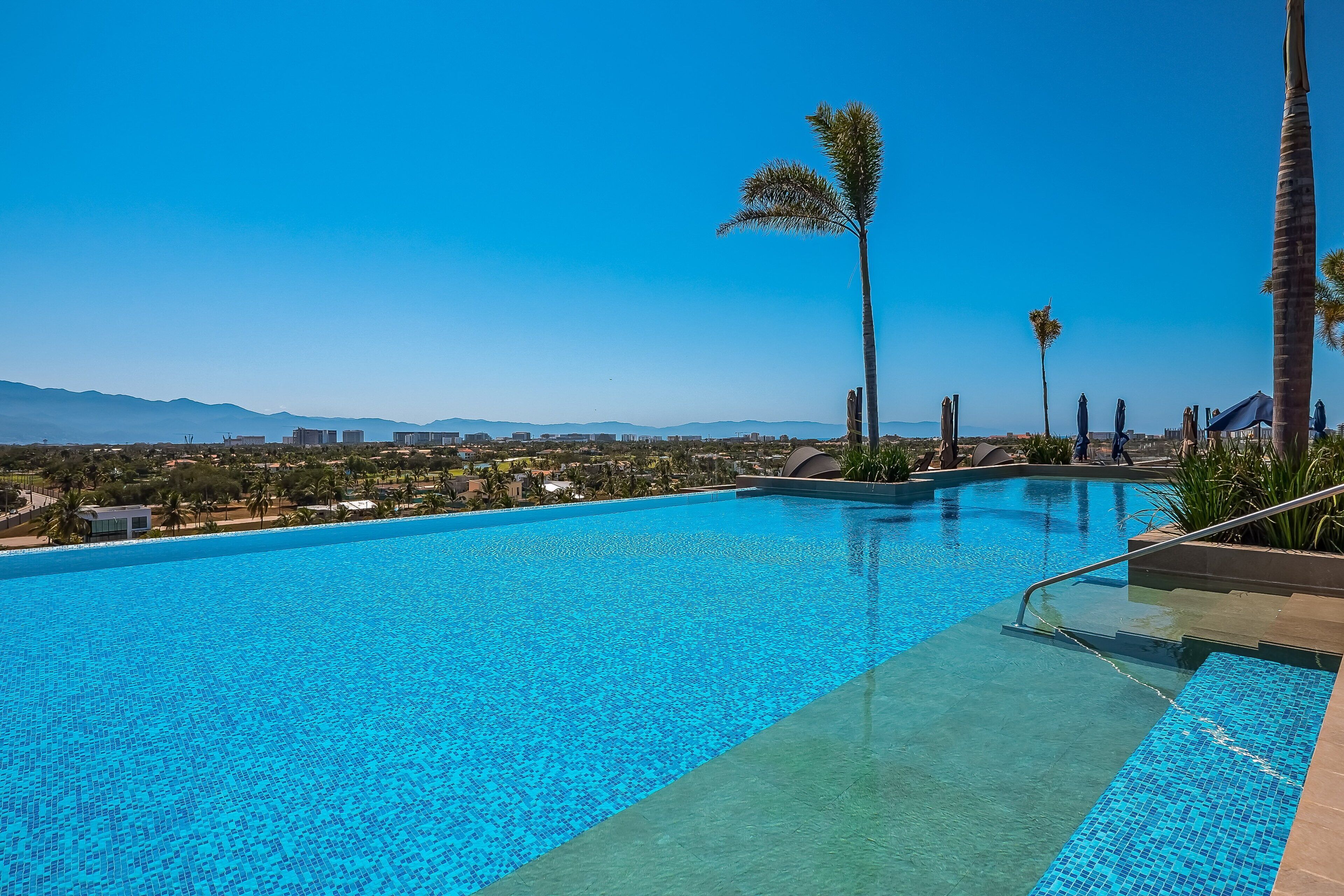 Ein Swimmingpool mit klarem, blauem Wasser, umgeben von Palmen und Blick auf entfernte Berge.