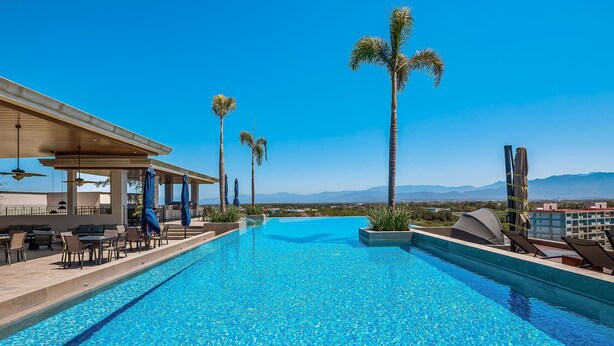 A clear blue pool with lounge chairs and umbrellas, surrounded by palm trees and a view of distant mountains. - D'toscana Residencial, PH #2 Apartment in Nuevo Vallarta (Nuevo Nayarit)