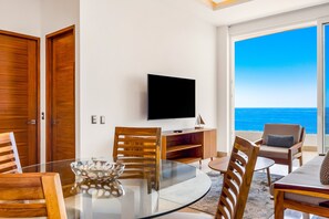 A modern living room with a glass table, wooden chairs, a flat-screen TV, and a view of the ocean. - 4th-floor Oceanfront Condo With Balcony, Outdoor Pool, Ocean Views & AC (Puerto Vallarta)