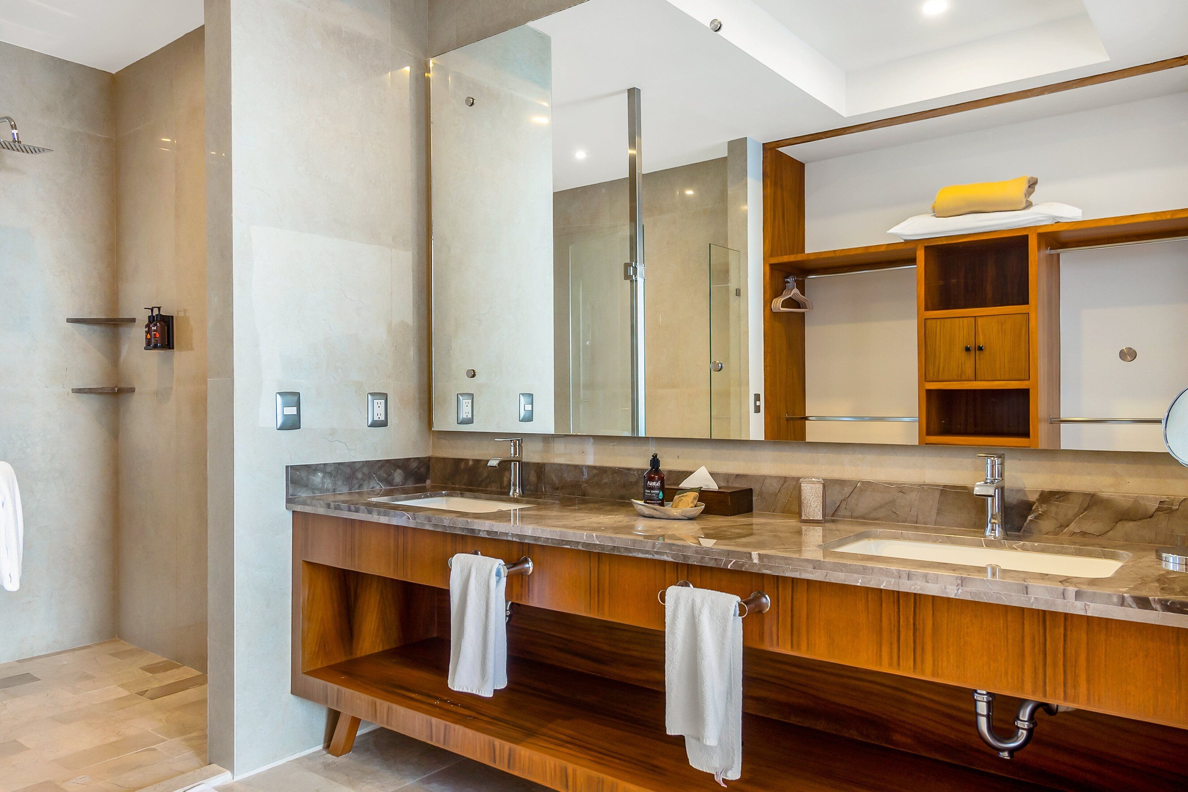 A modern bathroom with two sinks, a large mirror, wooden cabinetry, and a shower area.