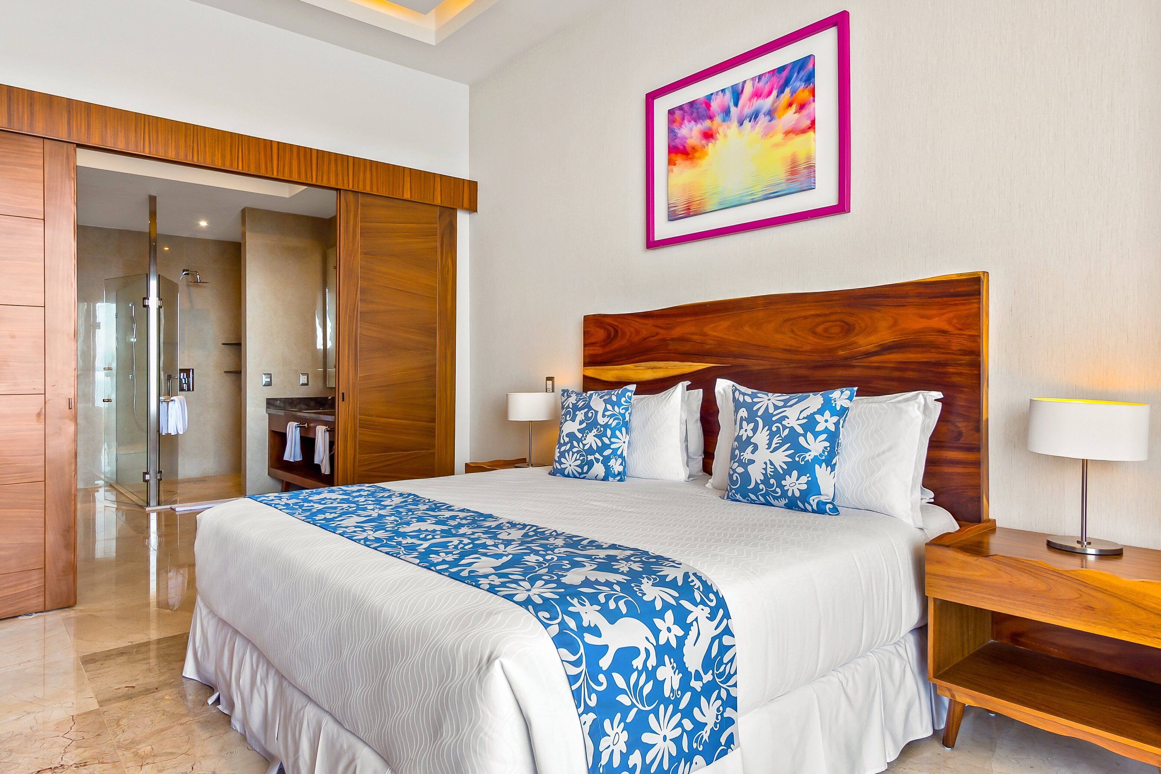 A hotel room with a large bed, wooden headboard, a glass shower, and a painting on the wall.