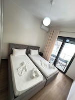 A hotel room with two beds, a ceiling fan, an air conditioning unit, a window with curtains, and a view of the outside.
