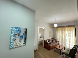 A living room with a sofa, coffee table, and a chair. There is a painting on the wall and a door leading to another room.