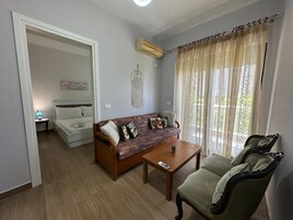 A living room with a sofa, a coffee table, and a chair. There is a bed in the background and a wall-mounted air conditioner.