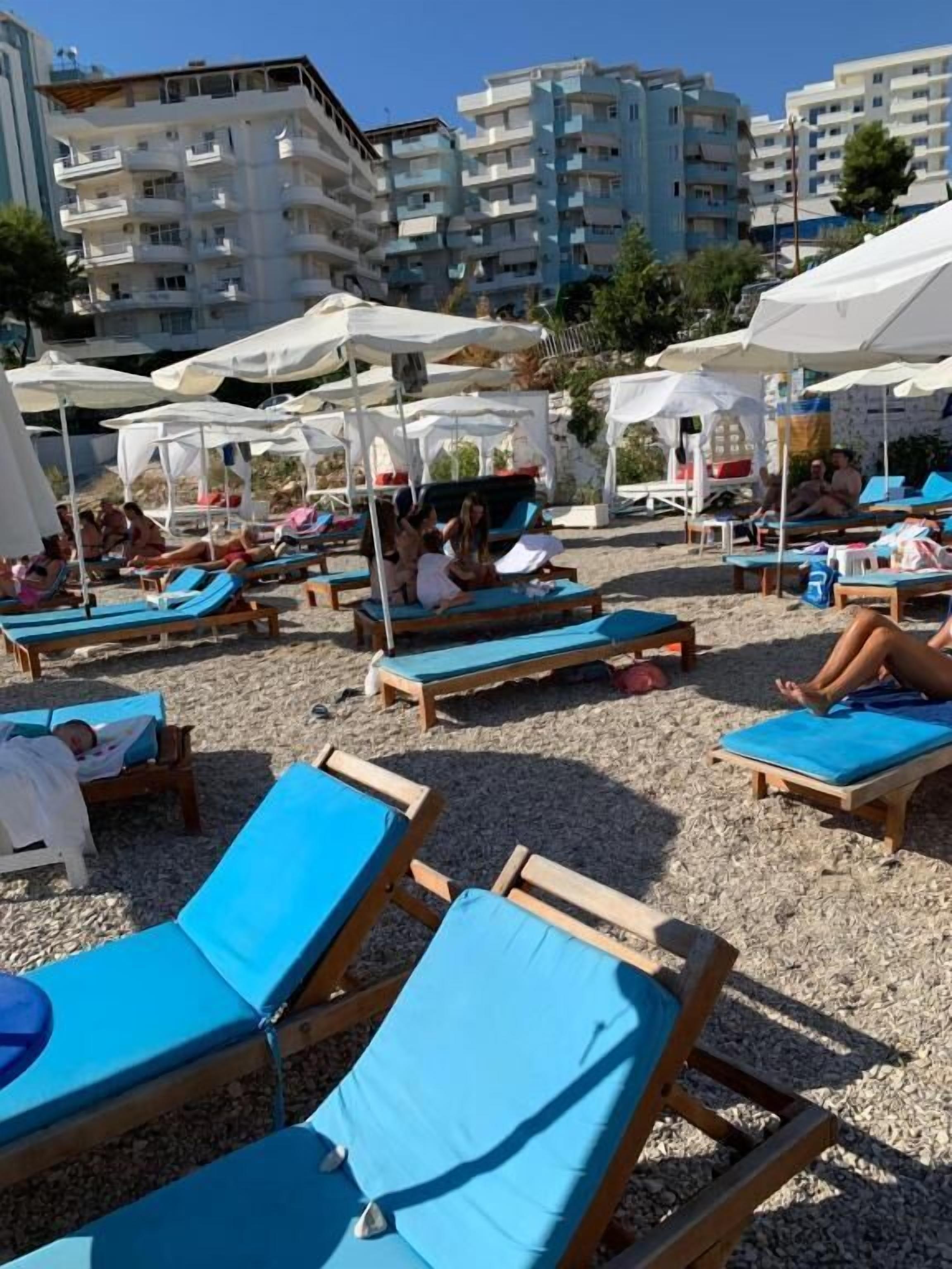 Ein Strand mit Liegestühlen und Sonnenschirmen, feiner Sand und Gebäude im Hintergrund.