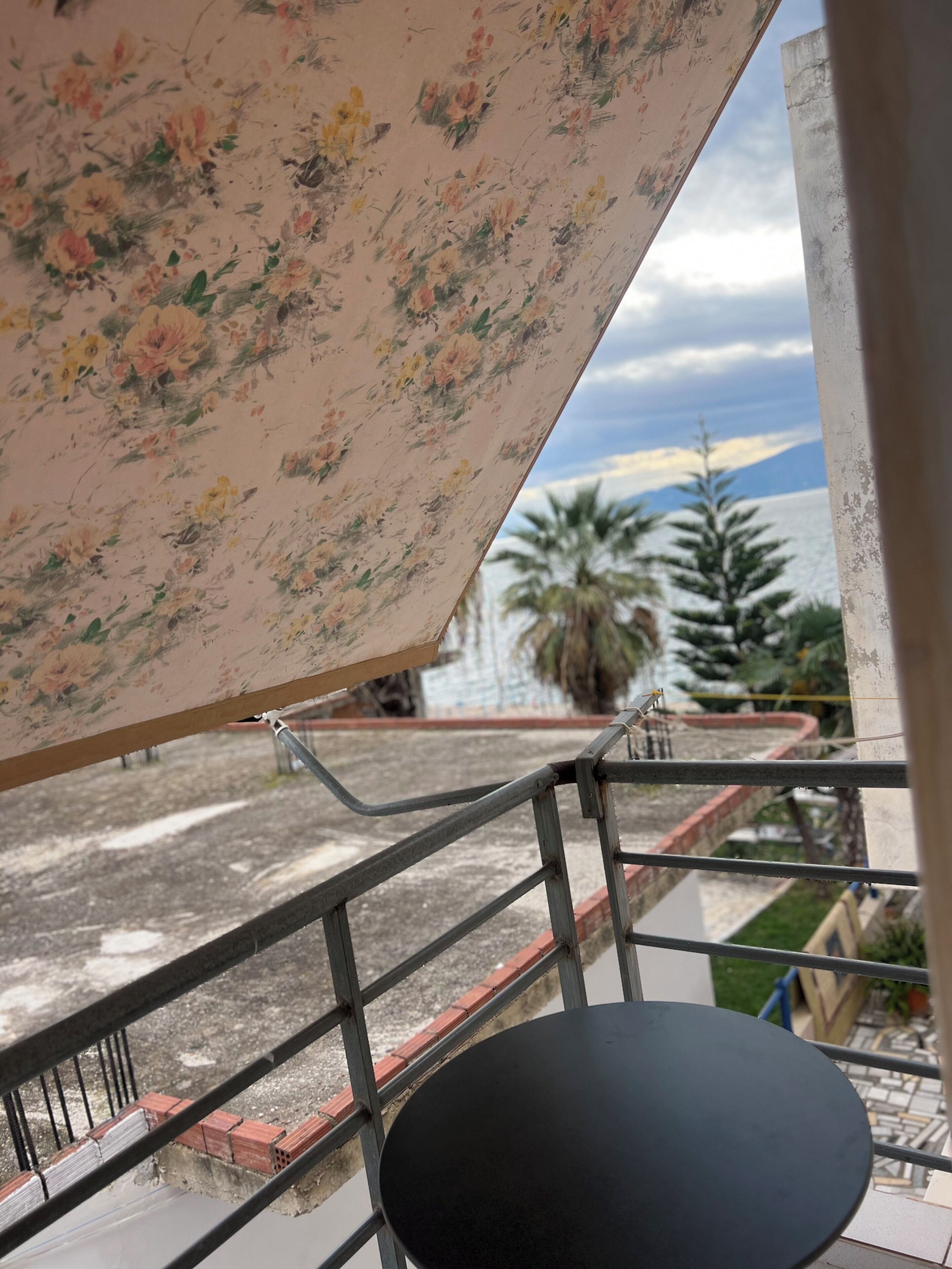 Un balcón con toldo estampado de flores, barandilla de metal y vista a un cuerpo de agua con palmeras y montañas al fondo.