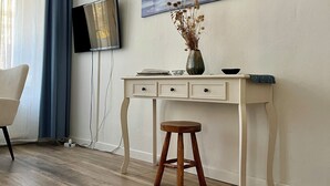 Una habitación con piso de madera, un escritorio blanco con cajones, un taburete de madera, un televisor de pantalla plana montado en la pared y un cuadro sobre el escritorio.