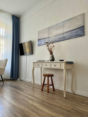 A room with a wooden floor, a white desk with drawers, a wooden stool, a flat-screen TV mounted on the wall, and a painting above the desk. - Cozy vacation apartment in a good location (Görlitz)