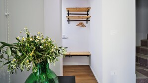 Een gang met een houten bankje, een groene vaas met bloemen en een wandrek met kledinghangers.