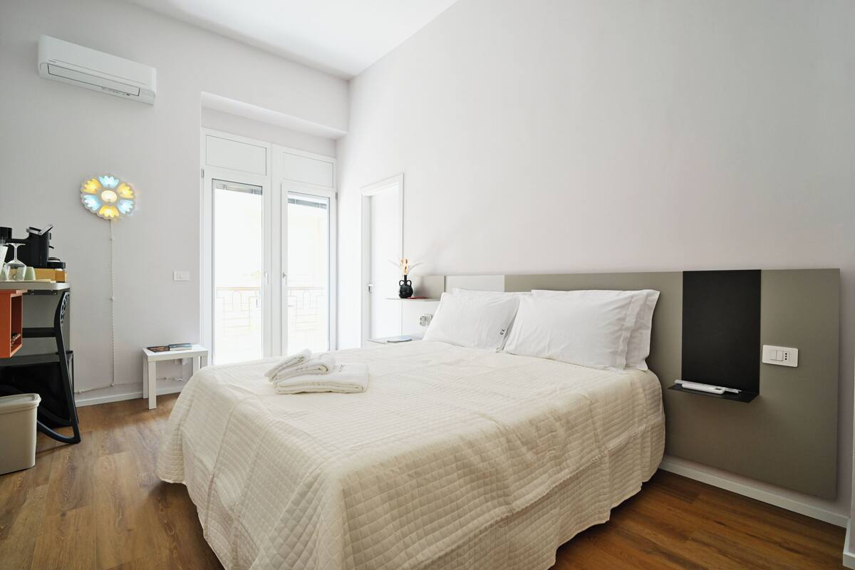 A bedroom with a bed, a bedside table, a nightstand, a wall-mounted shelf, and an air conditioning unit.