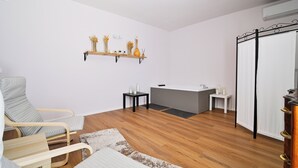 Een moderne kamer met een houten vloer, een grijze stoel, een klein tafeltje, een witte bank, een plank met decoratieve objecten, en een badruimte met een wastafel en spiegel.
