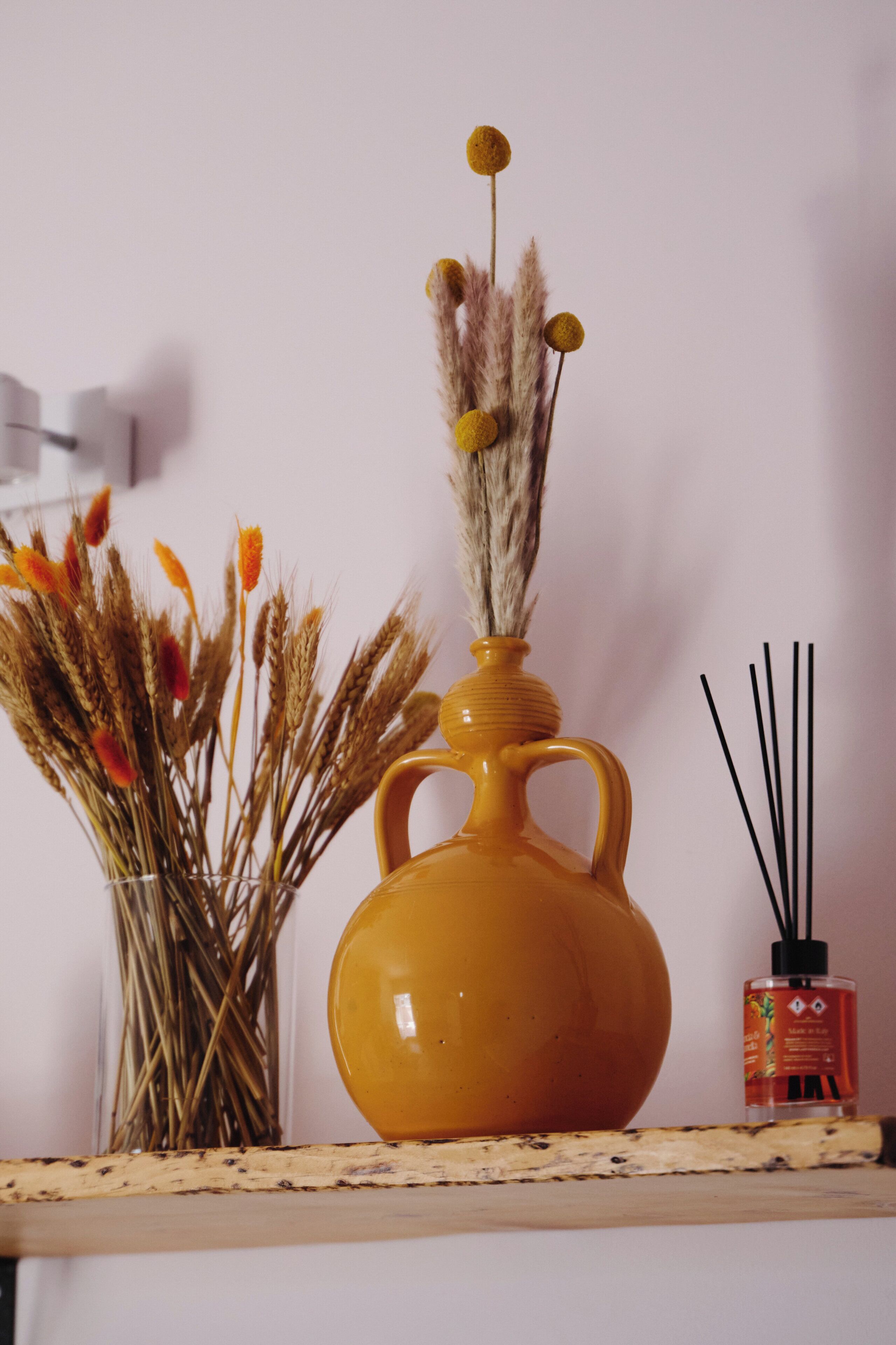 Un vase jaune contenant des plantes séchées et un flacon de diffuseur à bâtonnets, le tout posé sur une étagère.