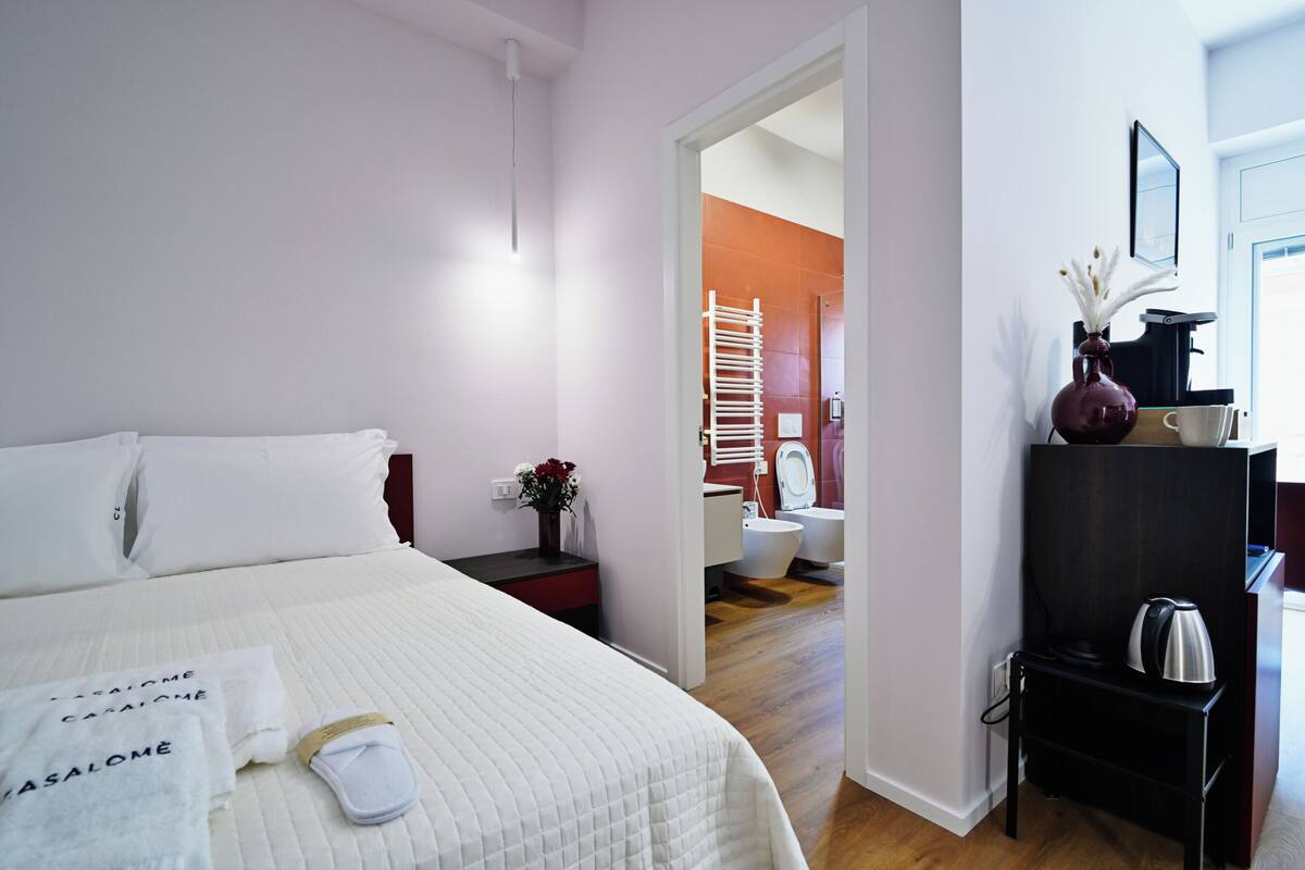 A hotel room with a bed, a nightstand with a vase of flowers, a coffee machine, and a view into the bathroom.