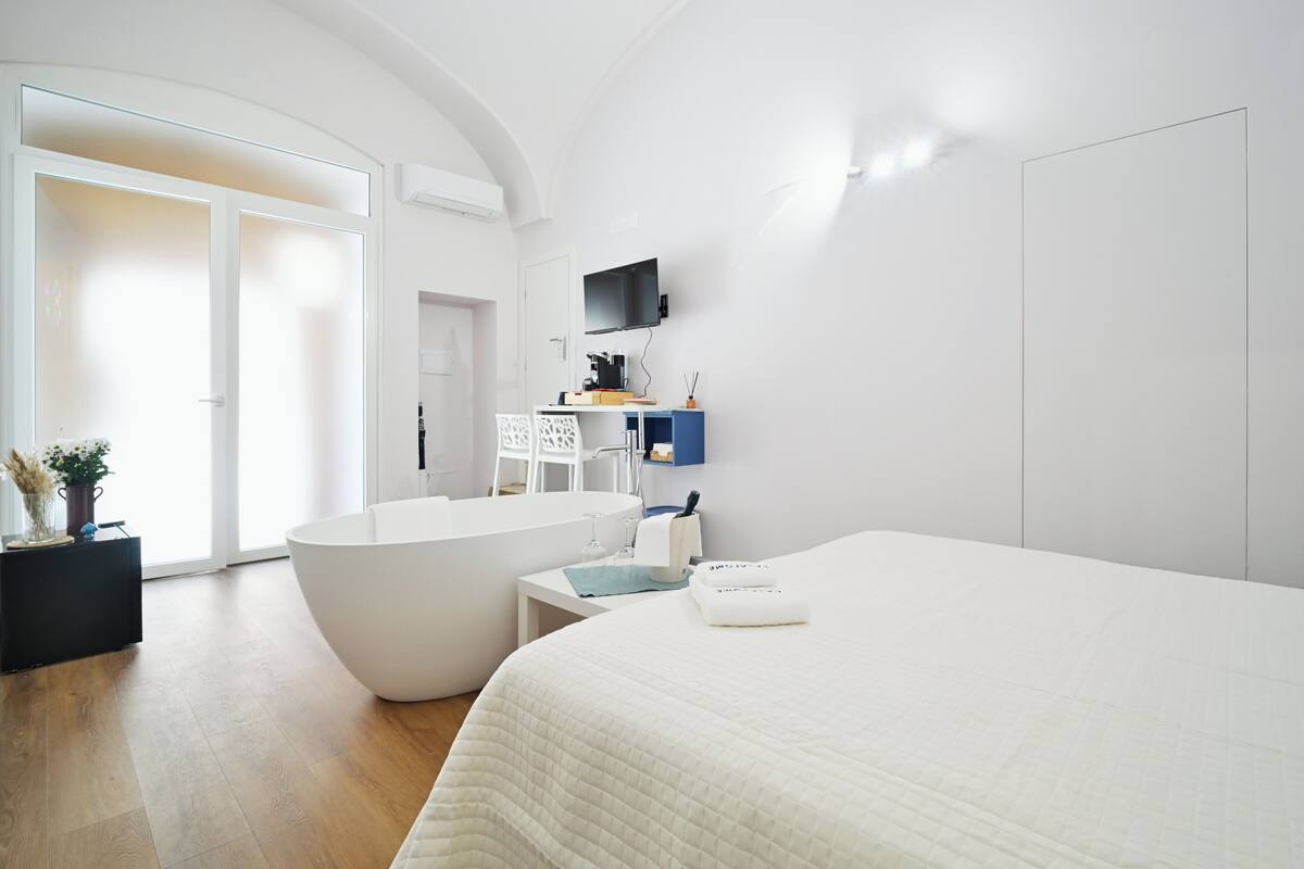 A modern hotel room with a large bed, a bathtub, a small table, and a TV mounted on the wall.