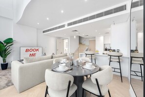 En moderne stue med sofa, spisebord, stole, et køkken i baggrunden og en trappe til et øvre plan.