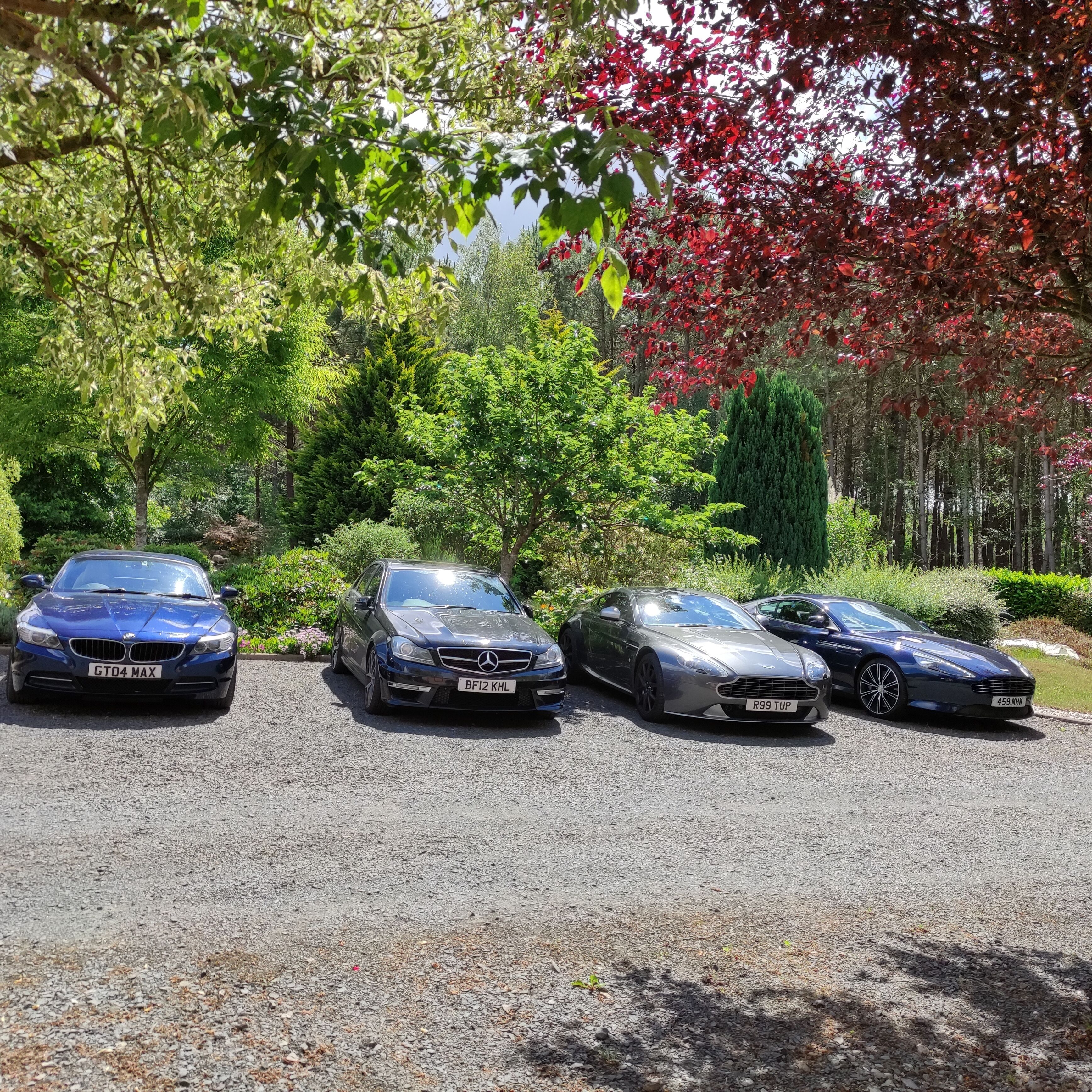 Three luxury cars parked on a gravel driveway surrounded by lush greenery and trees.