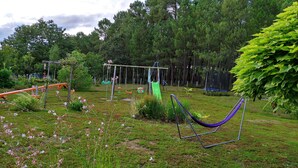 Een speeltuin met schommels, een glijbaan en een trampoline in een grasveld, met bomen op de achtergrond.