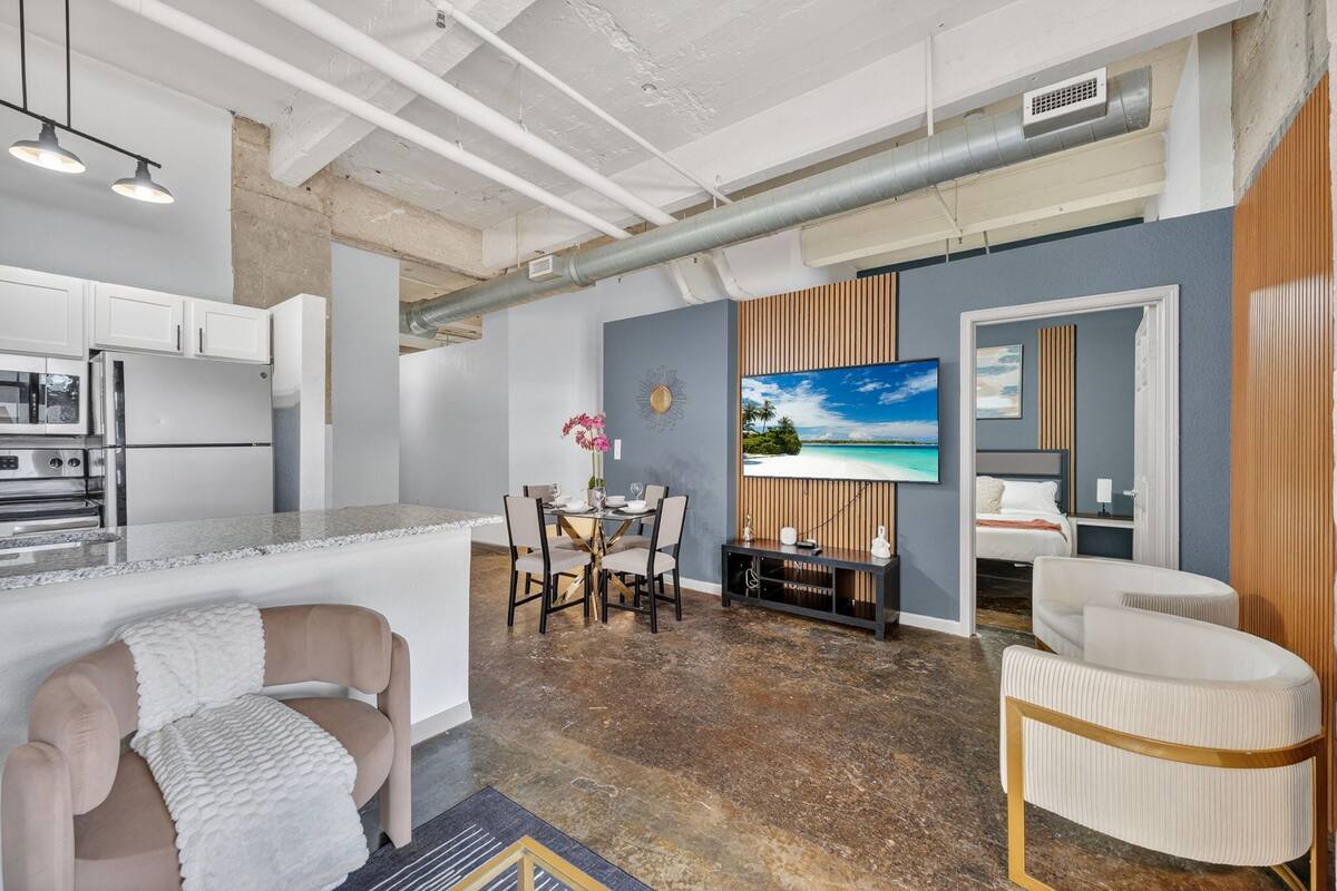 A modern kitchen with white cabinets, a dining area with a table and chairs, a TV mounted on a wall, and a bedroom with a bed and a window.