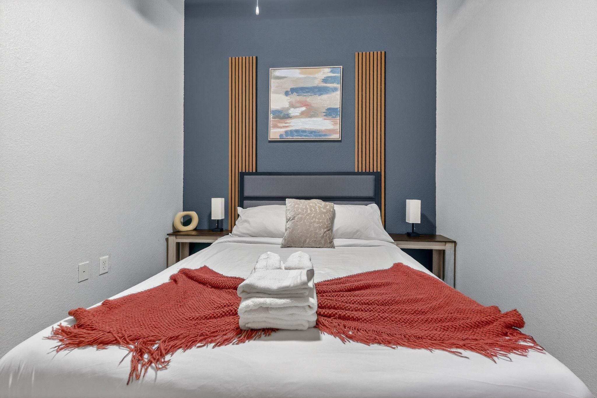 A neatly made bed with a red blanket, a stack of white towels, and a painting above it.
