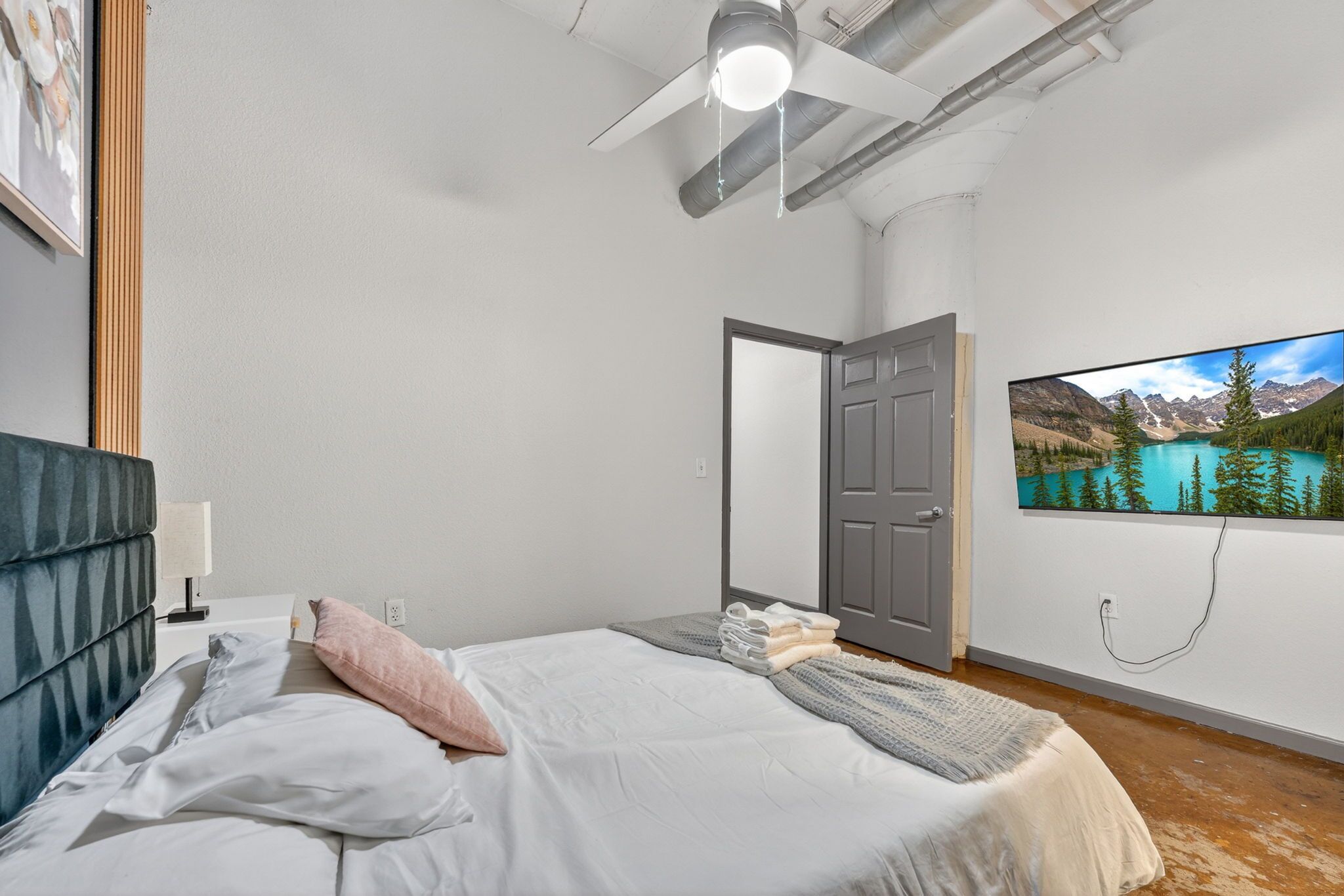 A bedroom with a bed, a ceiling fan, a door, and a wall-mounted TV displaying a mountain landscape.
