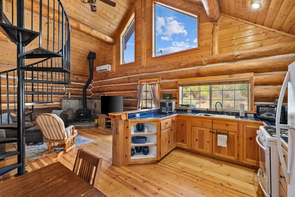 A log cabin interior with a spiral staircase, a fireplace, a kitchen with wooden cabinets, and a large window offering a view of the outdoors.