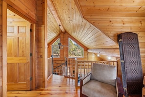 A wooden cabin interior with a loft bedroom, a staircase, and a view of the outdoors.