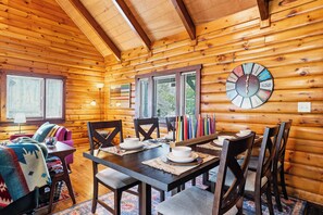 Interior de una cabaña de madera con una mesa de comedor para cuatro, una zona de estar acogedora y un reloj de pared.
