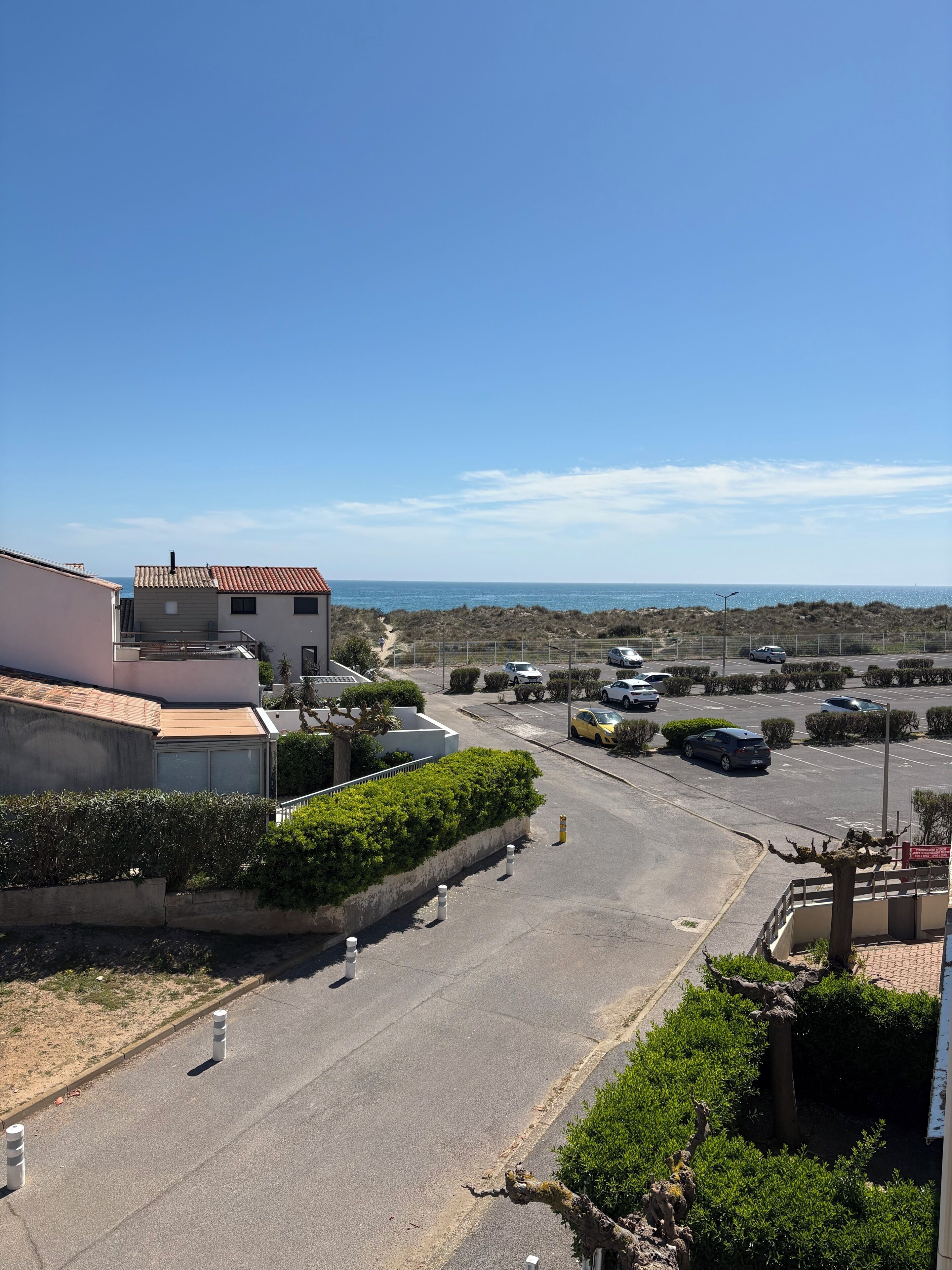 A residential street with parked cars, a clear sky, and a view of the sea in the distance.
