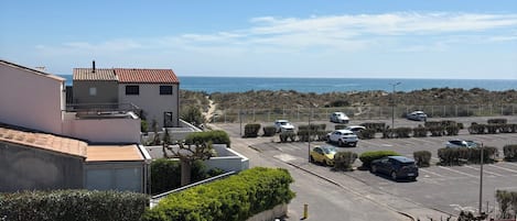 A residential street with parked cars, a clear sky, and a view of the sea in the distance.