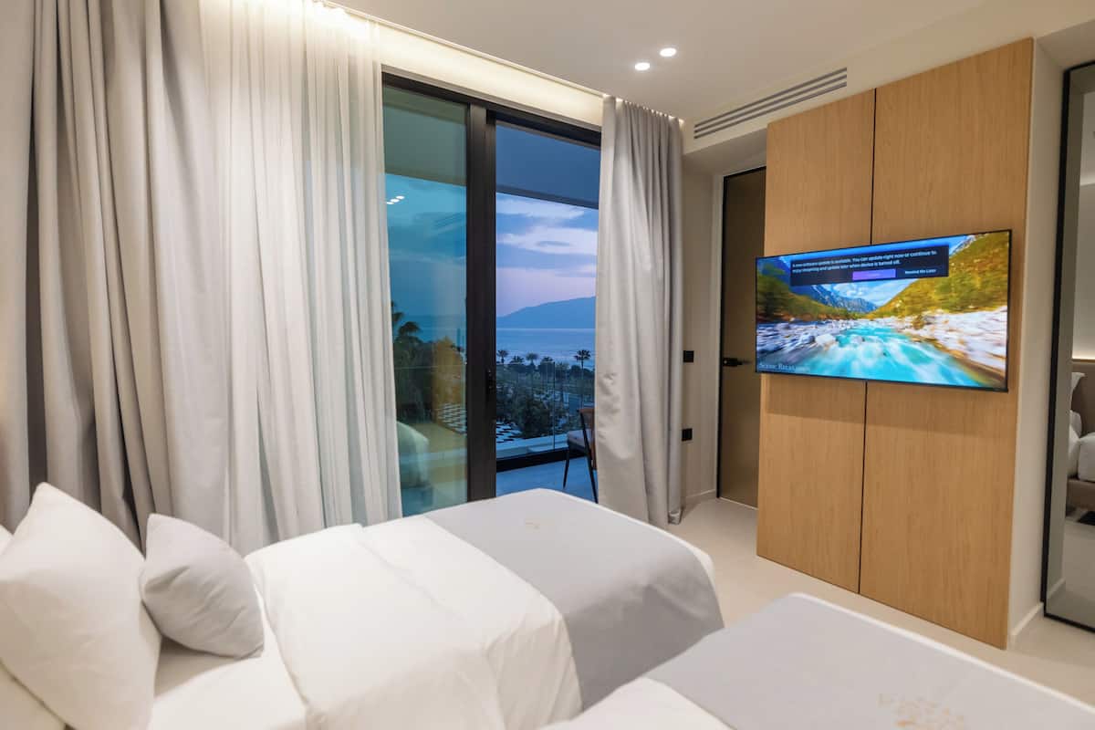 A modern hotel room with a large TV, a bed, a sofa, and a balcony with a view of the sea.