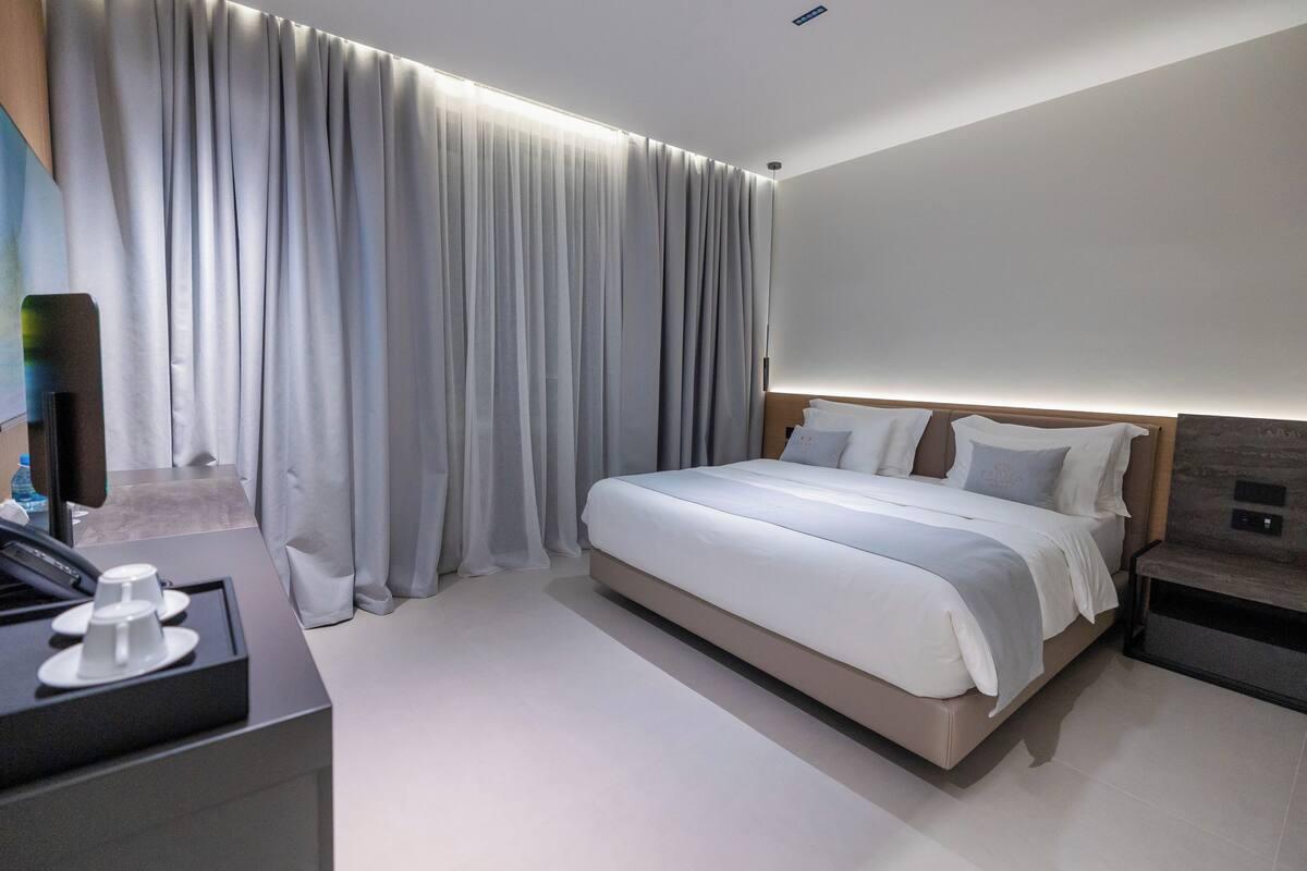 A modern hotel room with a large bed, a bedside table, a TV, and a coffee machine.