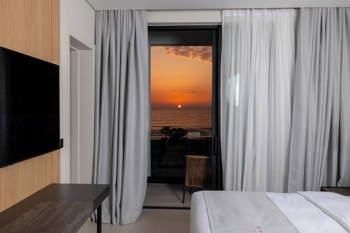 A hotel room with a bed, a TV mounted on the wall, a balcony with curtains, and a view of the sunset.